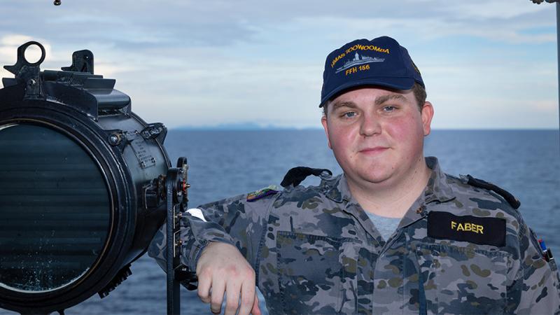 A military member stands with a device and the sea behind him. 