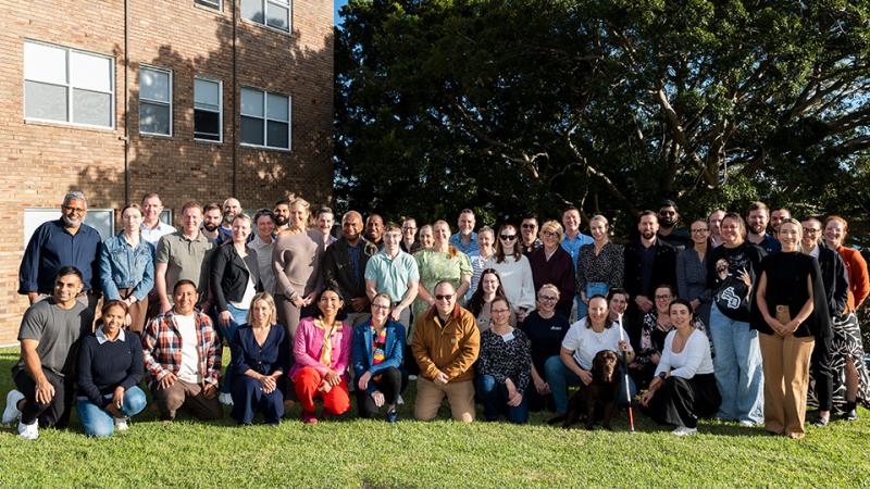 A group of about 50 people in casual clothing standing outside on the grass in front of a brick building and trees.