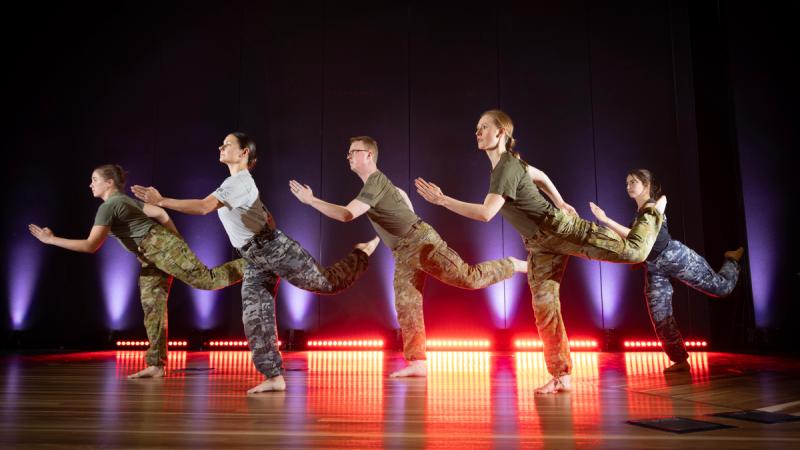 Five people stand barefoot on a stage in T-shirts and camouflage pants, arms and legs raised in the same exaggerated pose.
