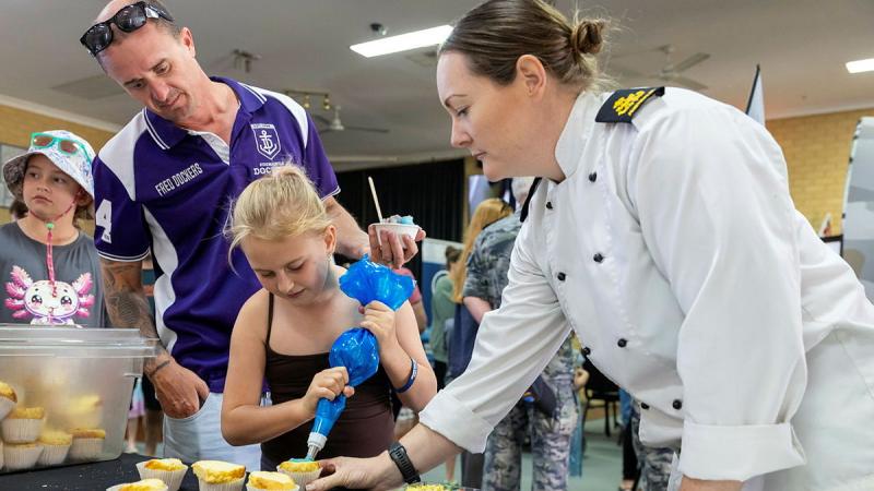 A Navy chef reaches to help a young girl who is decorating a cupcake with icing.
