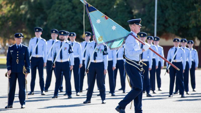 People on formal Air Force uniform stand in parade formation while one member walks in front of the group carrying a flag.