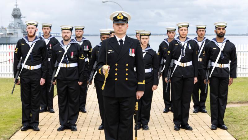 A man in formal Navy uniform stands to attention holding a sword, with a group of Navy personnel standing in uniform positions behind him.