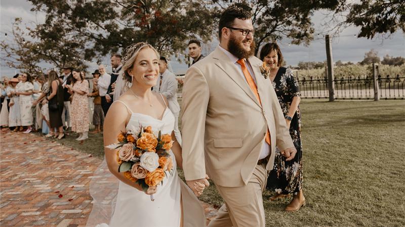 A man and woman walk down an outdoor wedding aisle in formal wear. 