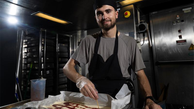 A man wearing an apron and flat-top chef’s hat in a commercial kitchen places a skewer into food in a baking tray.