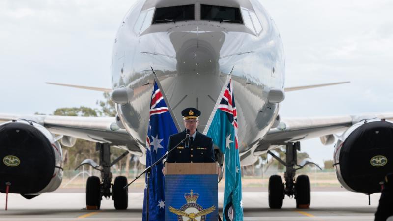 A man in Air Force uniform stands behind a lectern with two flags and a large plane behind him.