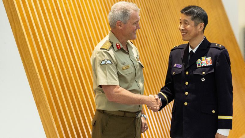Two men in uniform smiling and shaking hands with a wood-panelled wall in the background.