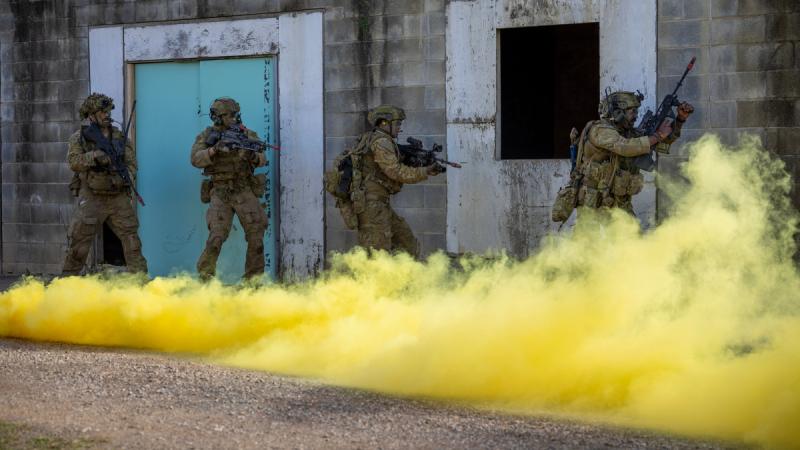 Four people in full camouflage uniform carrying guns move close to the wall of a cinderblock building, yellow smoke billowing in front of them.