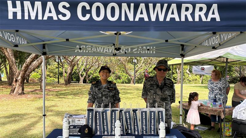 Two people in camouflage uniforms standing behind a table under an HMAS Coonawarra gazebo at a market stall.