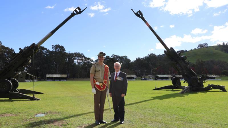 Two men stand on a grassed field in front of two large artillery cannons, one wearing a military uniform and the other a suit with medals.