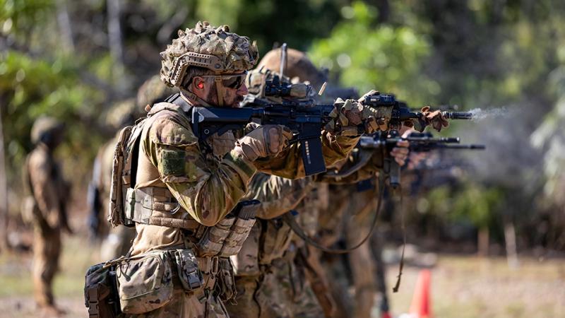 People wearing camouflage uniforms, helmets and glasses stand in a line firing weapons.