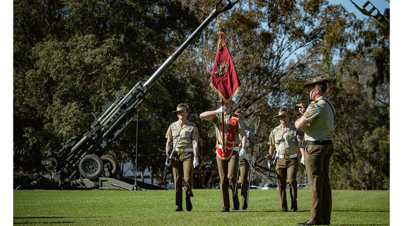 Military personnel with a red banner march past a large stationary weapon outside.