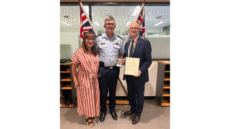 Two men and a woman stand before two flags with an award.