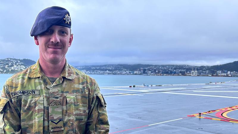 A man in camouflage uniform and a beret stands near water with a hilly coastline behind him.