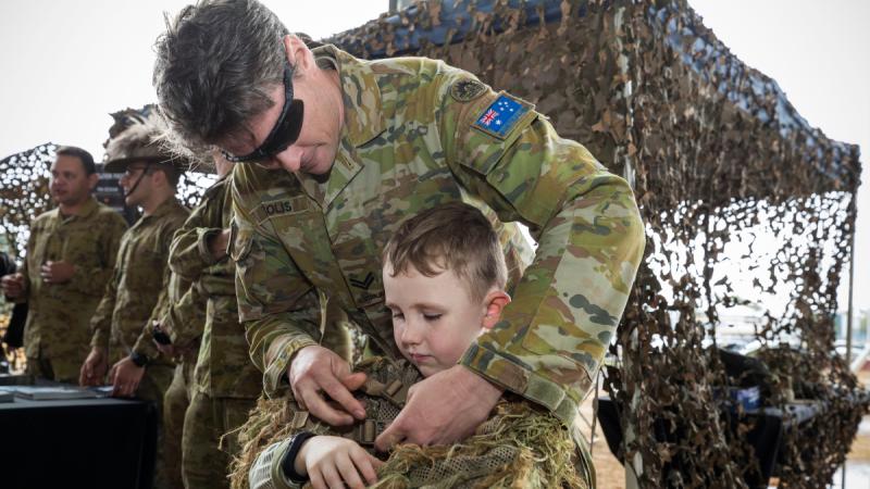A man in Army uniform helps a young boy dress in camouflage. 