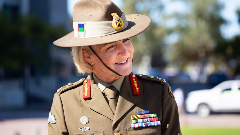 A woman in formal Army uniform, including a broad-brimmed hat, tilting her head and smiling outdoors.