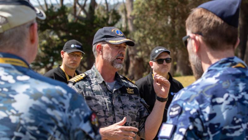 A man in Navy uniform speaks to other uniformed personnel gathered around.
