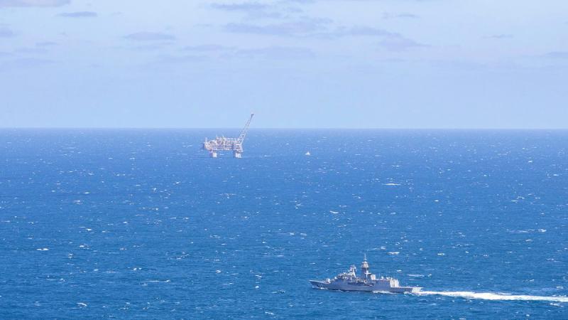 A ship sails close to an oil platform in the middle of the ocean on a clear, sunny day.