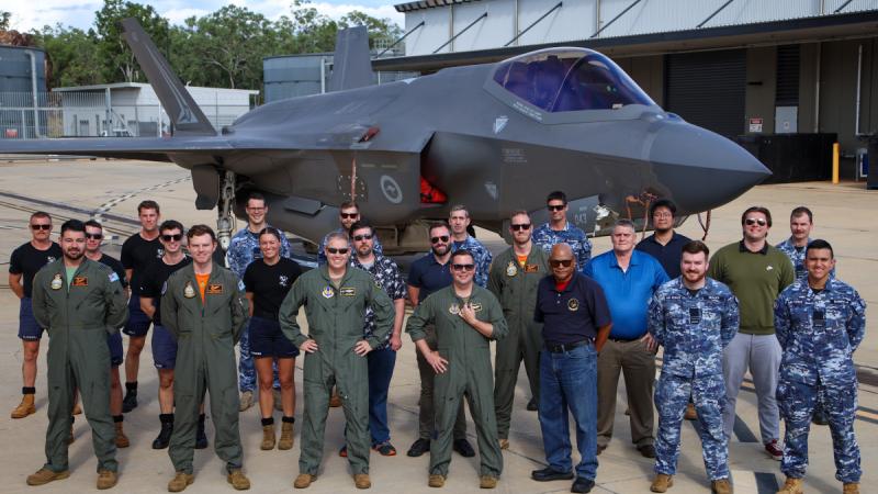 Numerous people, some in flight suits, stand in front of a jet aircraft.