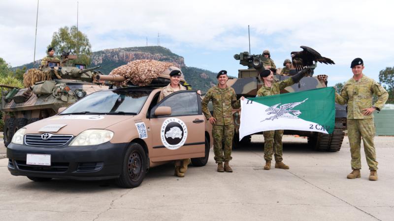 Squadron Leader Jessica Aldred stands behind the open passenger door of 'Tankette' beside three members of the army holding 2nd Cavalry Regiment's unit flag. Behind the ADF members is two M1A2 Main Battle Tanks.