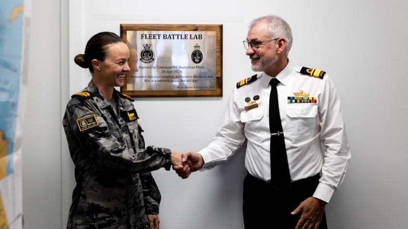 Two people in uniform shaking hands in front of a plaque. 