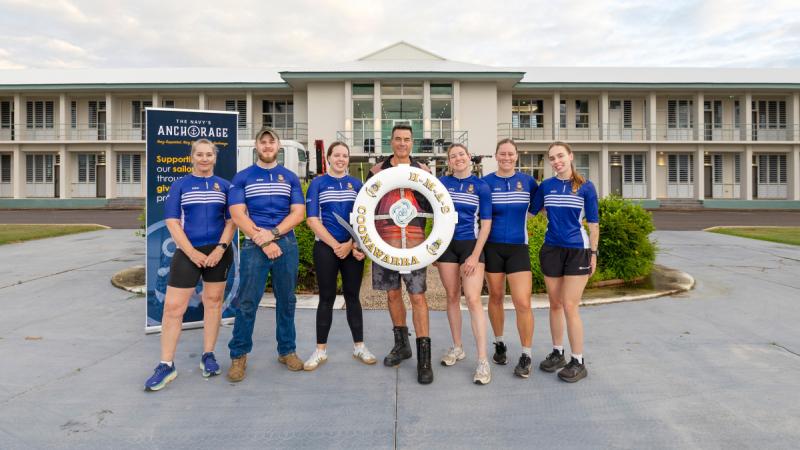 A group of seven people, most wearing racing shirts, stand in front of a large building, with the person in the centre holding a lifebuoy marked HMAS Coonawarra.