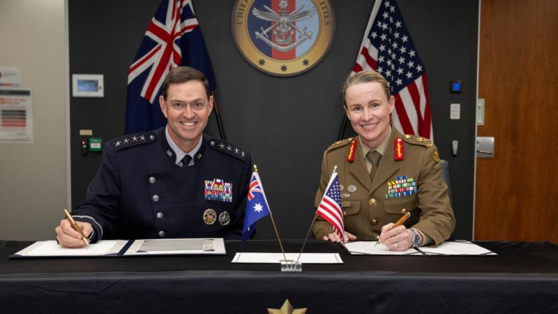 Two people in uniform sit at a table with small Australian and US flags, signing documents, with larger flags behind them.