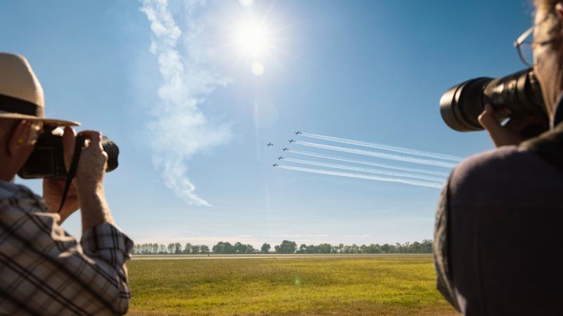 Two people holding cameras photograph aircraft flying in formation over a grassy airfield.