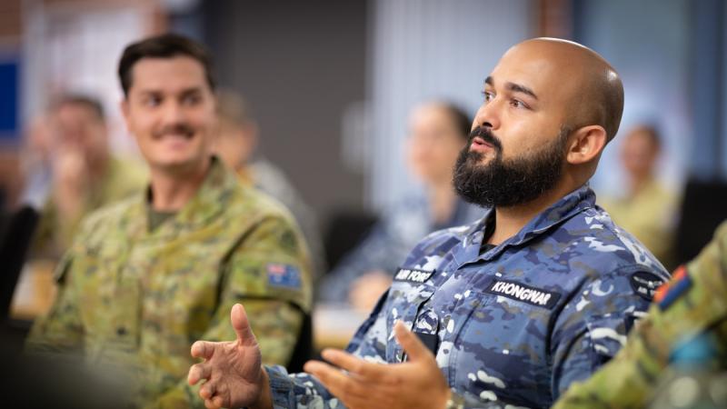 A man in a blue camouflage uniform gestures while seated, with another person in green camouflage seated beside him.