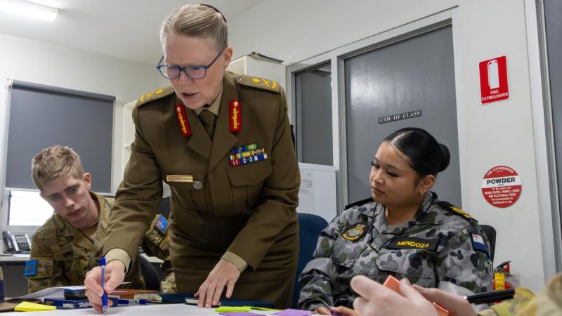An Army Major General leans over a table instructing two cadets who look on.