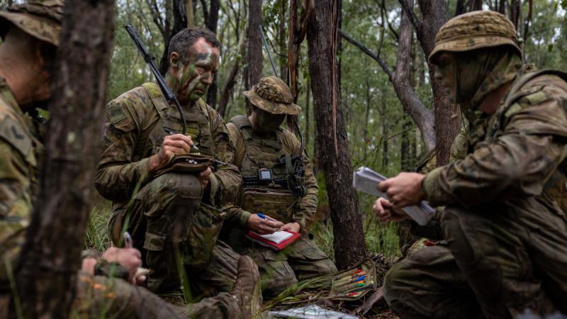 People in camouflage uniforms and face paint crouching in a forest and writing in notepads.