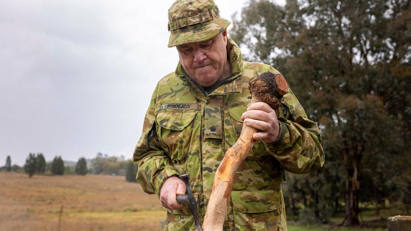 An Army member uses a small axe to shape a piece of wood.