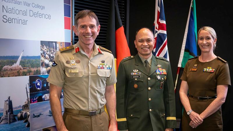Three people indifferent uniforms stand in front of various flags.