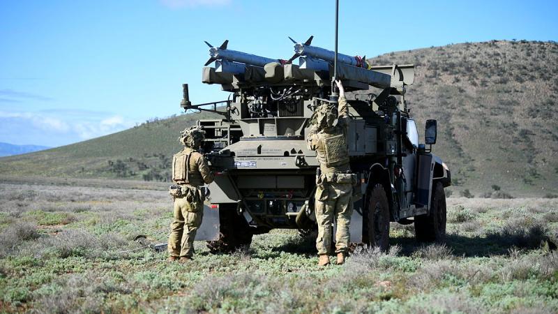 Soldiers stand behind an Army vehicle in the middle of grassy terrain.