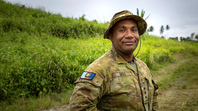 An Army member in uniform stands in a lush green setting.
