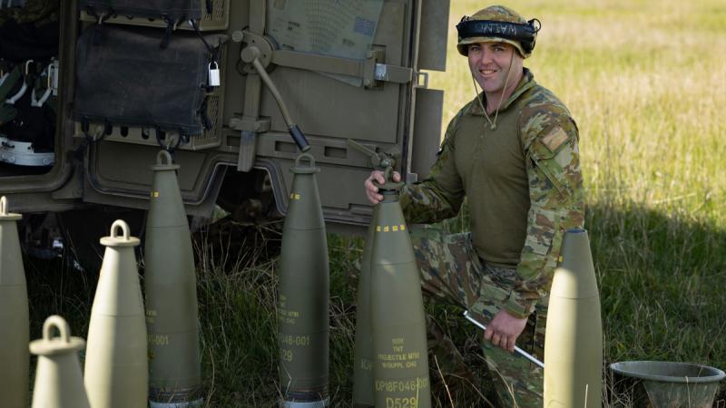 A soldier with large rounds squatting down in front of an Army vehicle.
