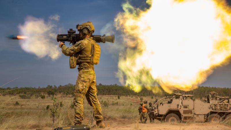 A person in camouflage fires a launcher while a projectile leaves the tube and a plume shoots from the rear, with others standing nearby.