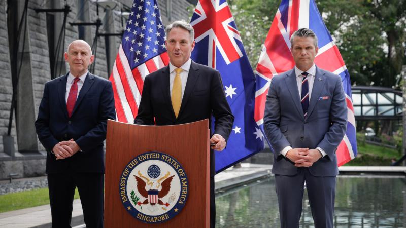Australia's Defence Minister speaks behind a lectern outside, with the United Kingdom and United States Defence Ministers on either side and three flags behind.