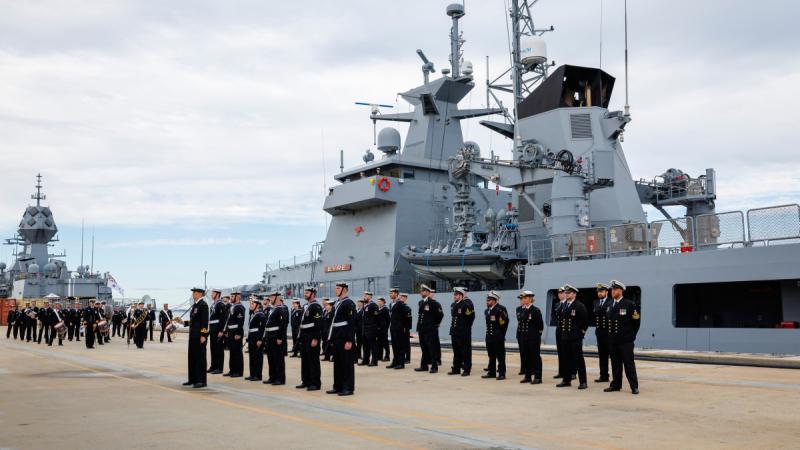Three rows of people in Navy uniform stand in formation in front of a naval ship as a marching band approaches.