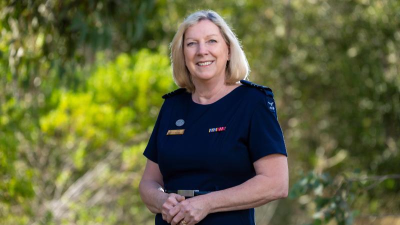 A woman in uniform stands outdoors, smiling, with greenery in the background.