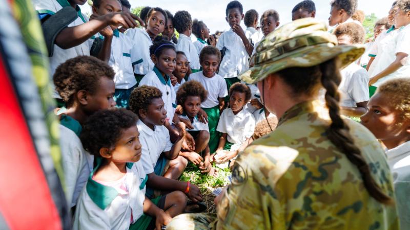 A woman in camouflage uniform sits on the grass surrounded by children wearing school uniforms.