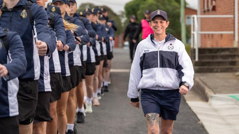 A person in sports uniform walks along while a line of people in sports uniform march along.