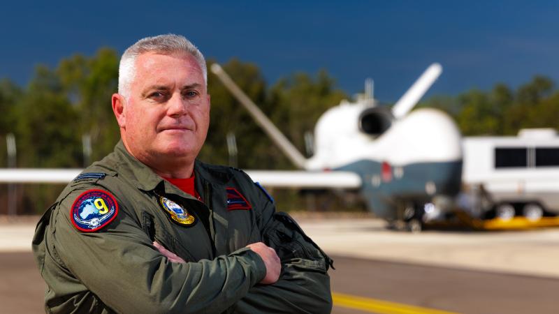 A man in military pilot gear stands with his arms crossed in front of an aircraft.