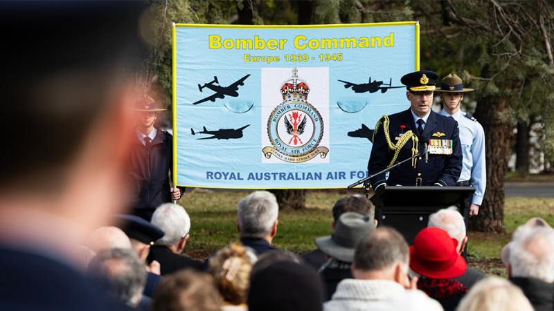 A uniformed man speaks at a lectern beside a Bomber Command banner as an audience faces him outdoors.