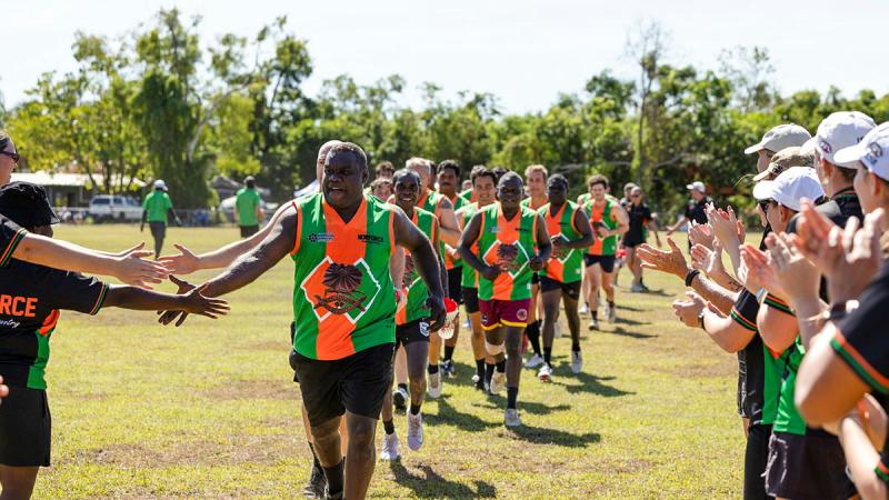 A team of football players in green and orange uniforms tap hands with each other in a line as they run across a football field.