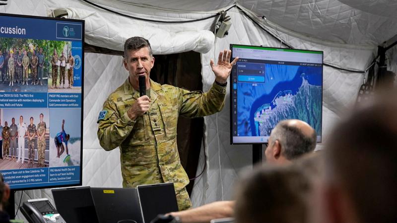 An Army member addresses a room of people with images on screens in the background.
