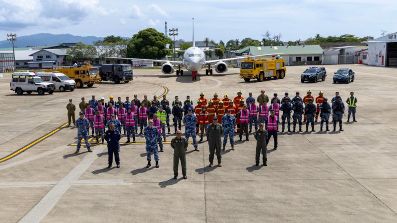 Around 50 people stand in a triangular formation in front of an aircraft and emergency vehicles.