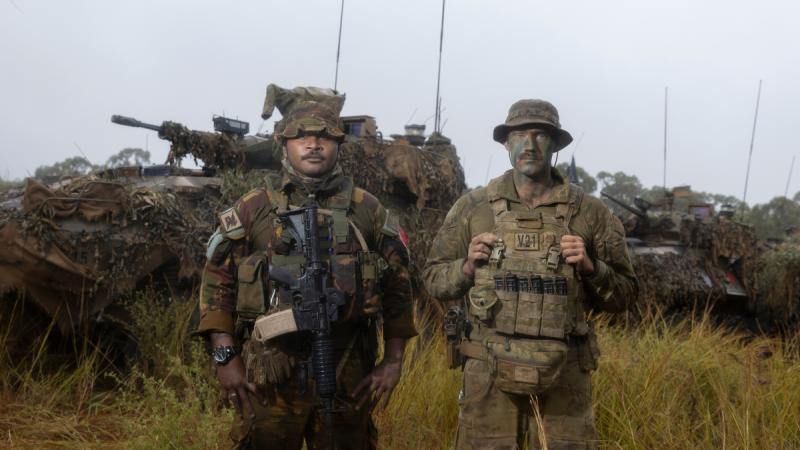 Two men in camouflage stand in tall grass near camouflaged armoured vehicles.
