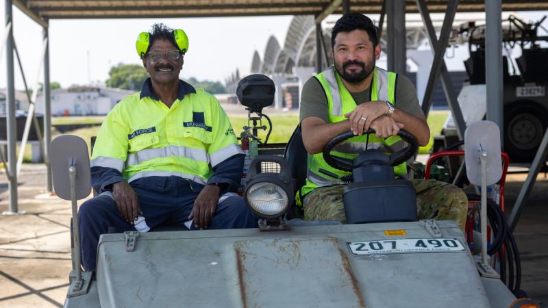 Two men in high-visibility clothing sit in a small vehicle, with aircraft hangars in the background.