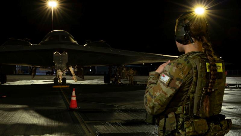 An Australian Air Force member stands on a runway, looking directly at a US stealth aircraft in low light.