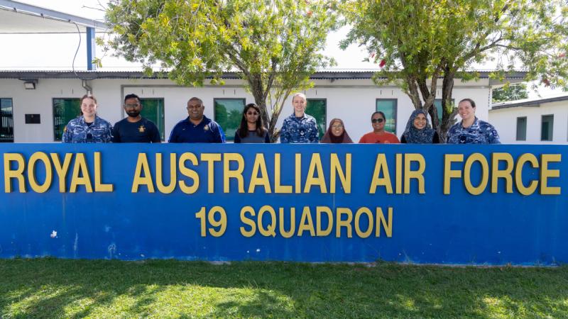 A group of people, some in uniform, stand behind a blue fence with the words ‘Royal Australian Air Force 19 Squadron’ on it in large yellow letters, with trees and a one-storey building behind them.
